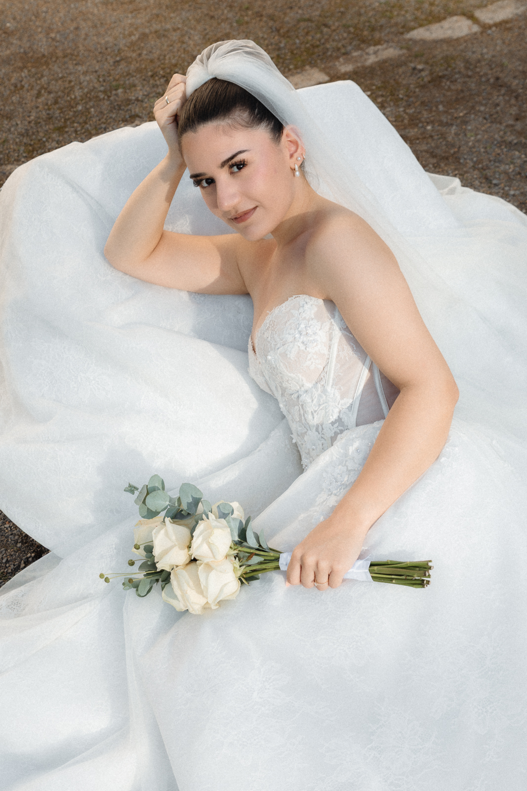 Braut sitzt in ihrem ausgebreiteten Kleid und blickt in die Kamera, in der Hand ein Strauß weißer Rosen, Aufnahme von oben