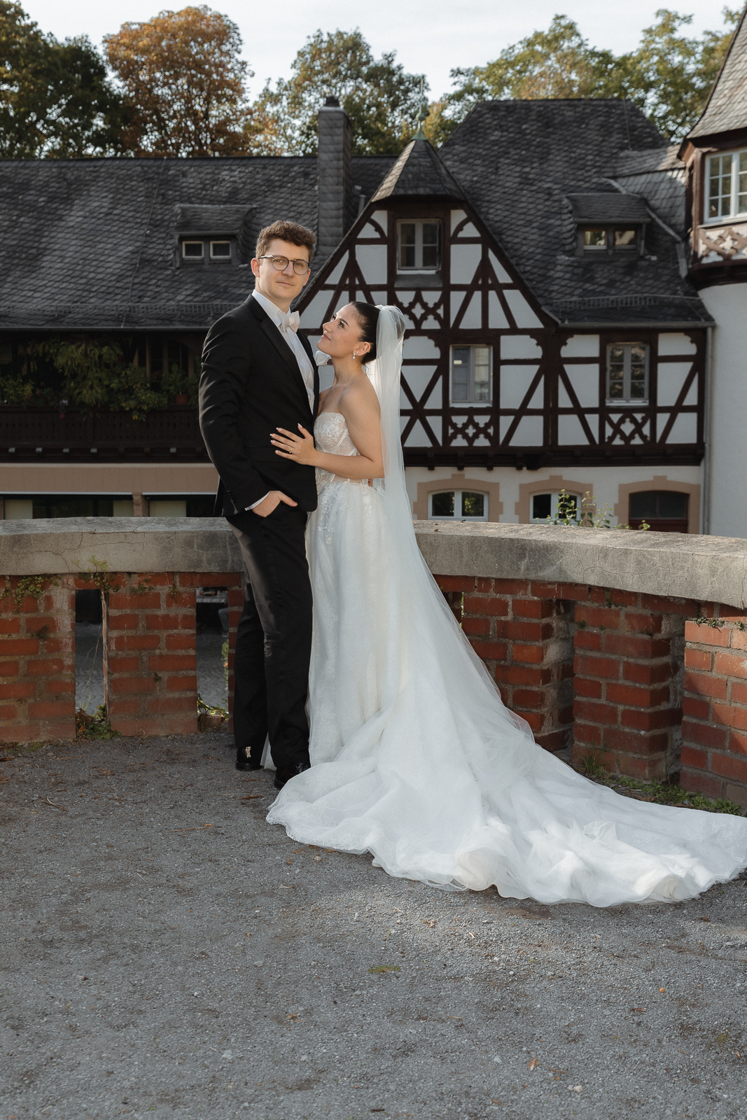 Brautpaar steht an einer Ziegelbrüstung vor einem Fachwerkhaus, die Braut blickt zum Bräutigam auf