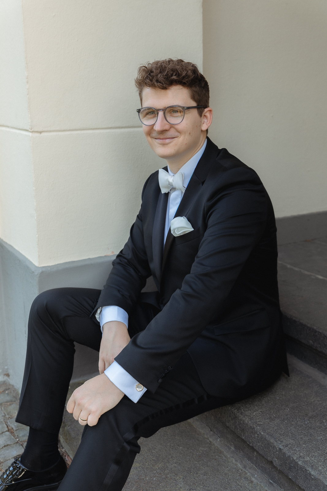 Bräutigam mit Brille und heller Fliege sitzt lächelnd auf einer Steinstufe