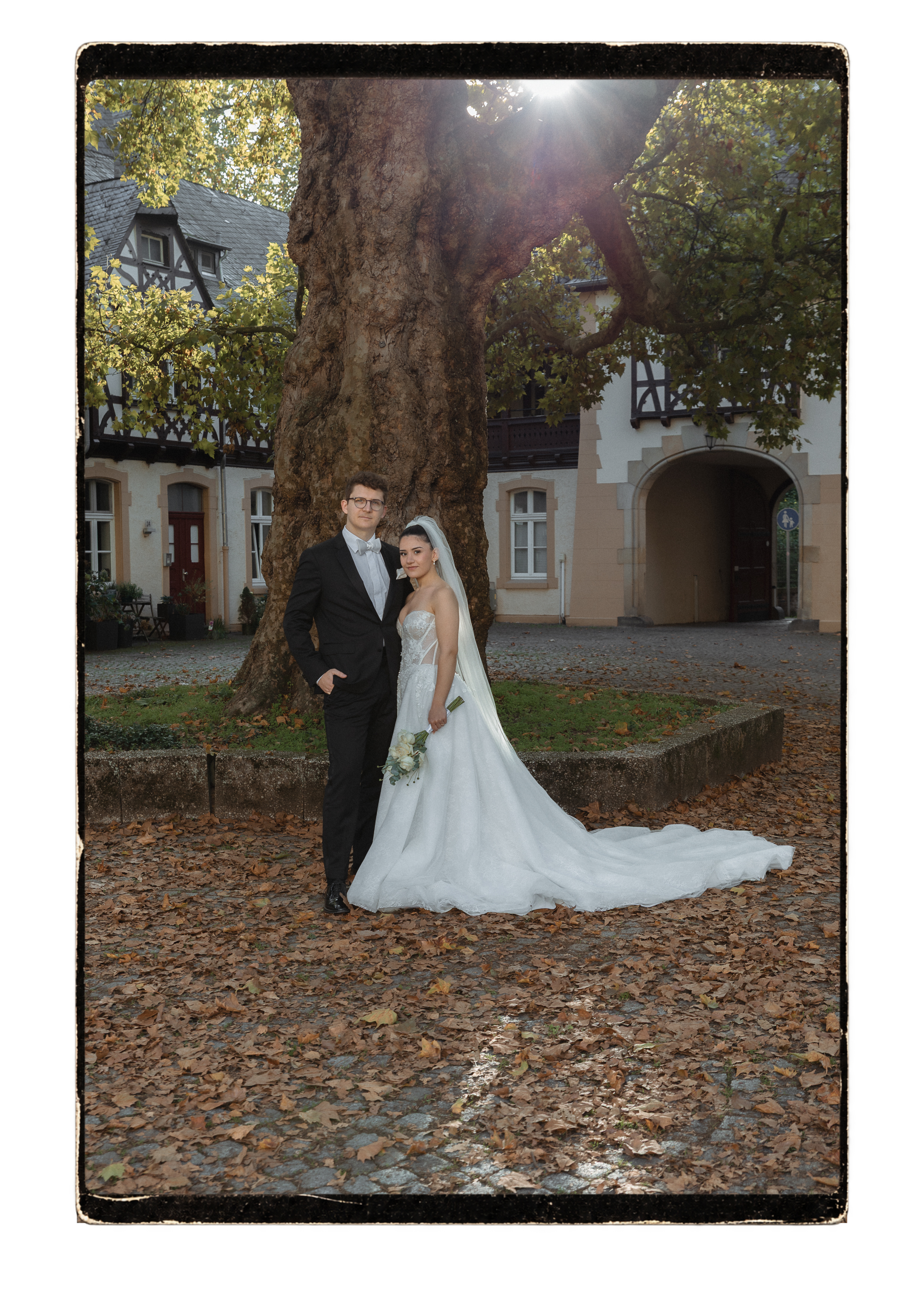 Brautpaar steht vor einem großen alten Baum im Innenhof eines Fachwerkensembles, Herbstlaub auf dem Boden, Aufnahme mit Filmrand