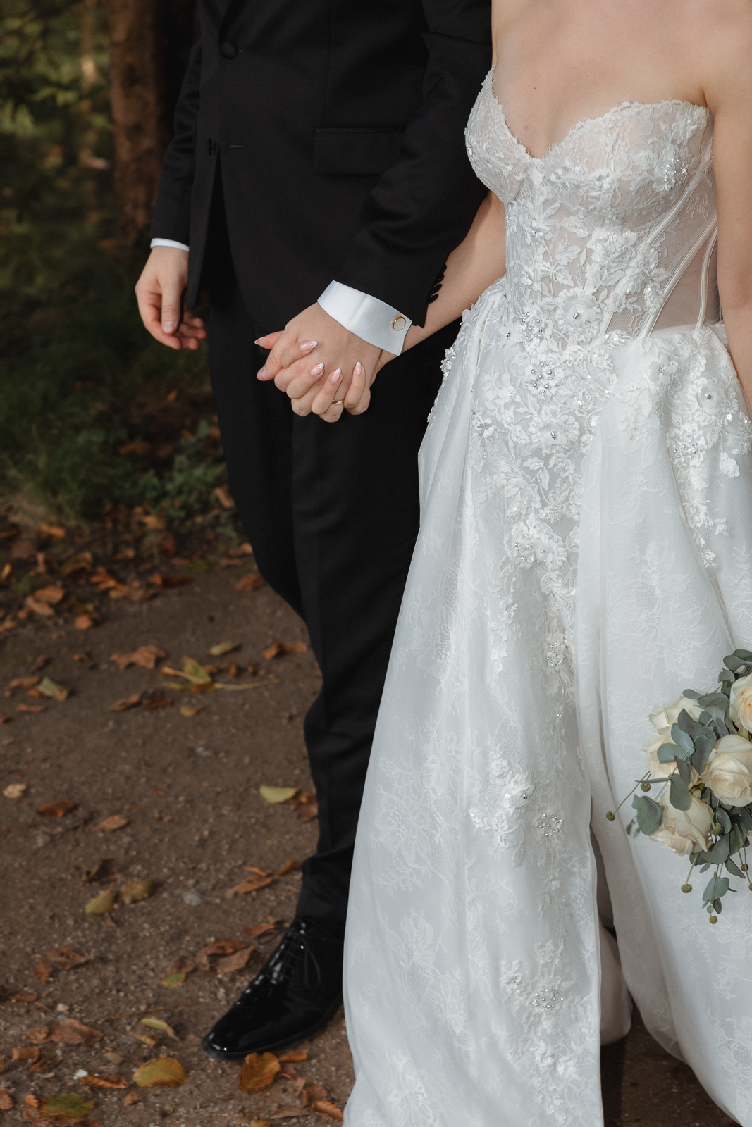 Detailaufnahme der ineinander gelegten Hände des Brautpaares, daneben das Spitzenkleid und der Brautstrauß