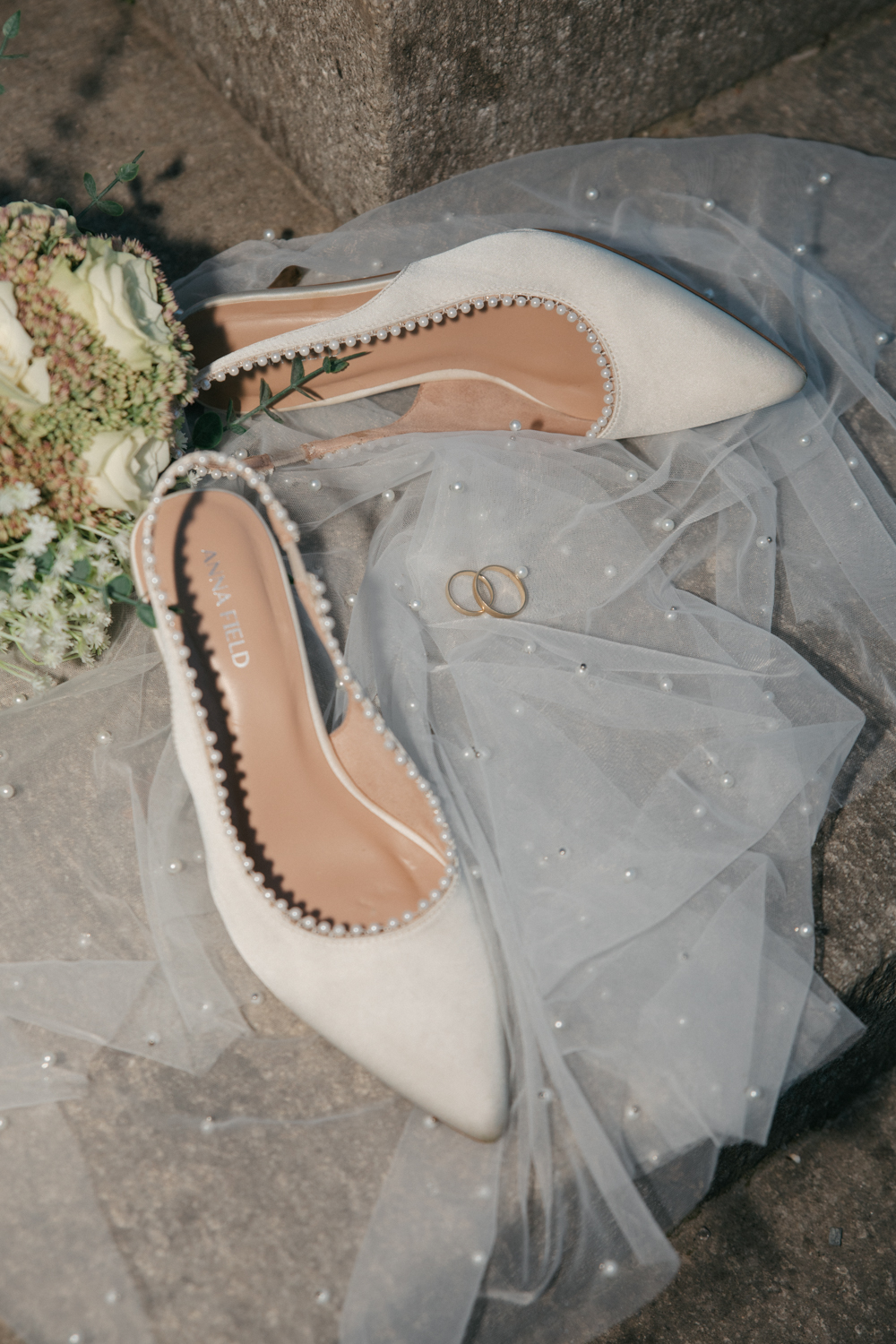 Brautschuhe mit Perlenbesatz, Eheringe und Brautstrauß auf einem perlenbestickten Schleier