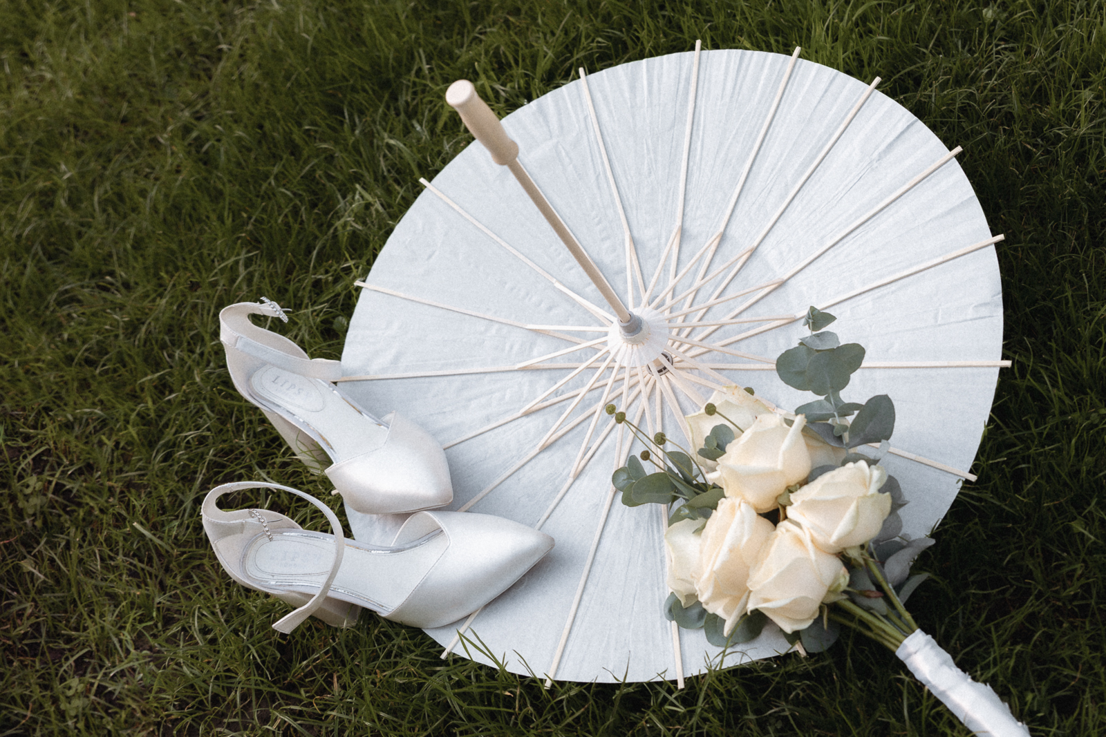 Weiße Brautschuhe, ein weißer Papierschirm und ein Brautstrauß aus weißen Rosen mit Eukalyptus auf dem Rasen