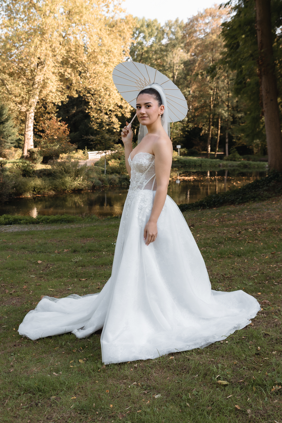 Braut im trägerlosen Spitzenkleid mit weißem Papierschirm vor einem Teich und herbstlichen Bäumen