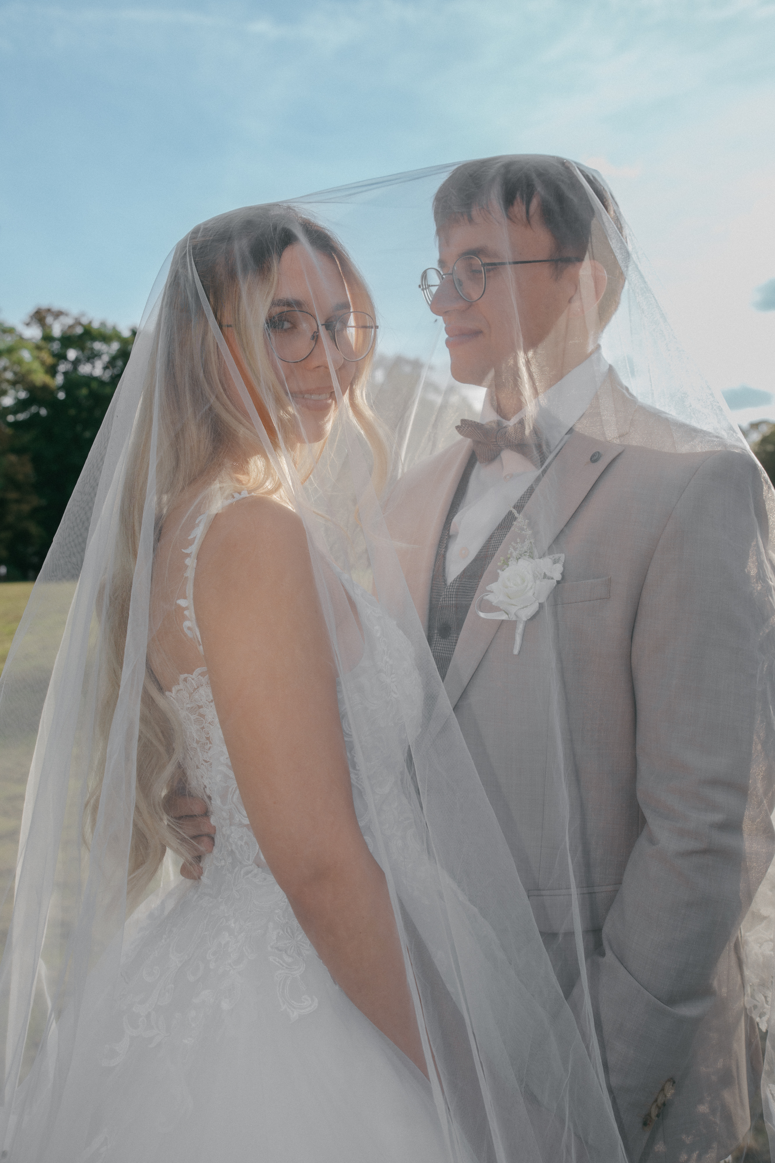 Braut mit Brille blickt unter dem gemeinsamen Schleier in die Kamera, der Bräutigam lächelt sie an